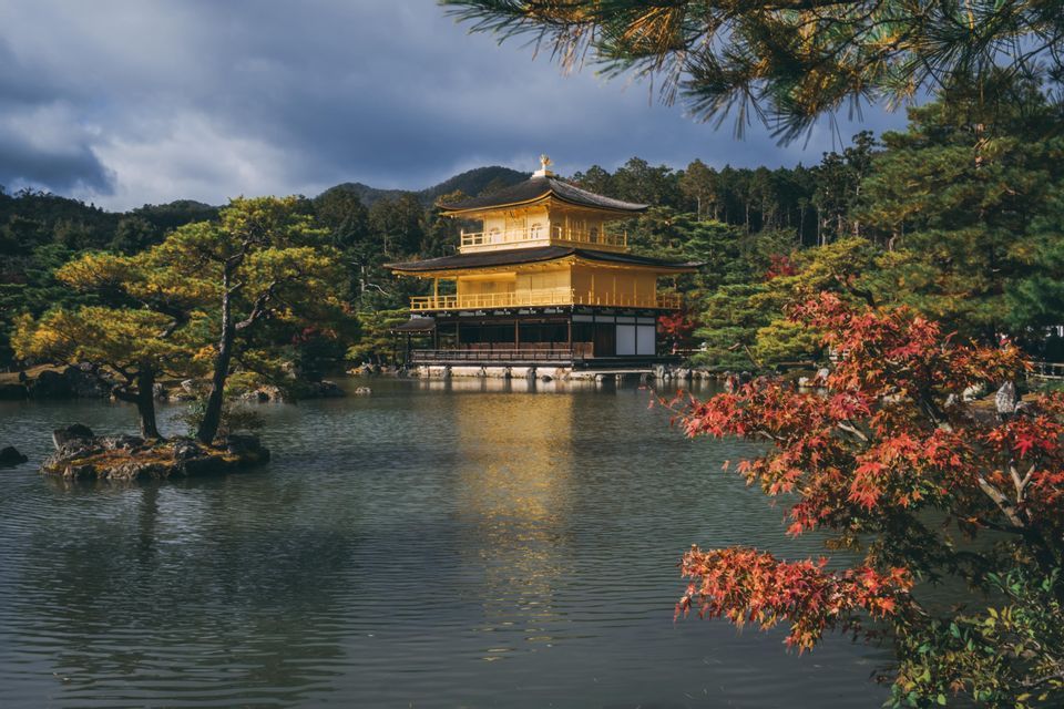 A golden Japanese temple stands by a reflective pond, surrounded by autumn trees and forested hills under a cloudy sky.