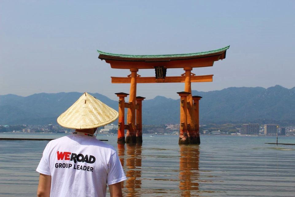Un chef de groupe WeRoad portant un chapeau conique contemple une grande porte torii orange se dressant dans l'eau.