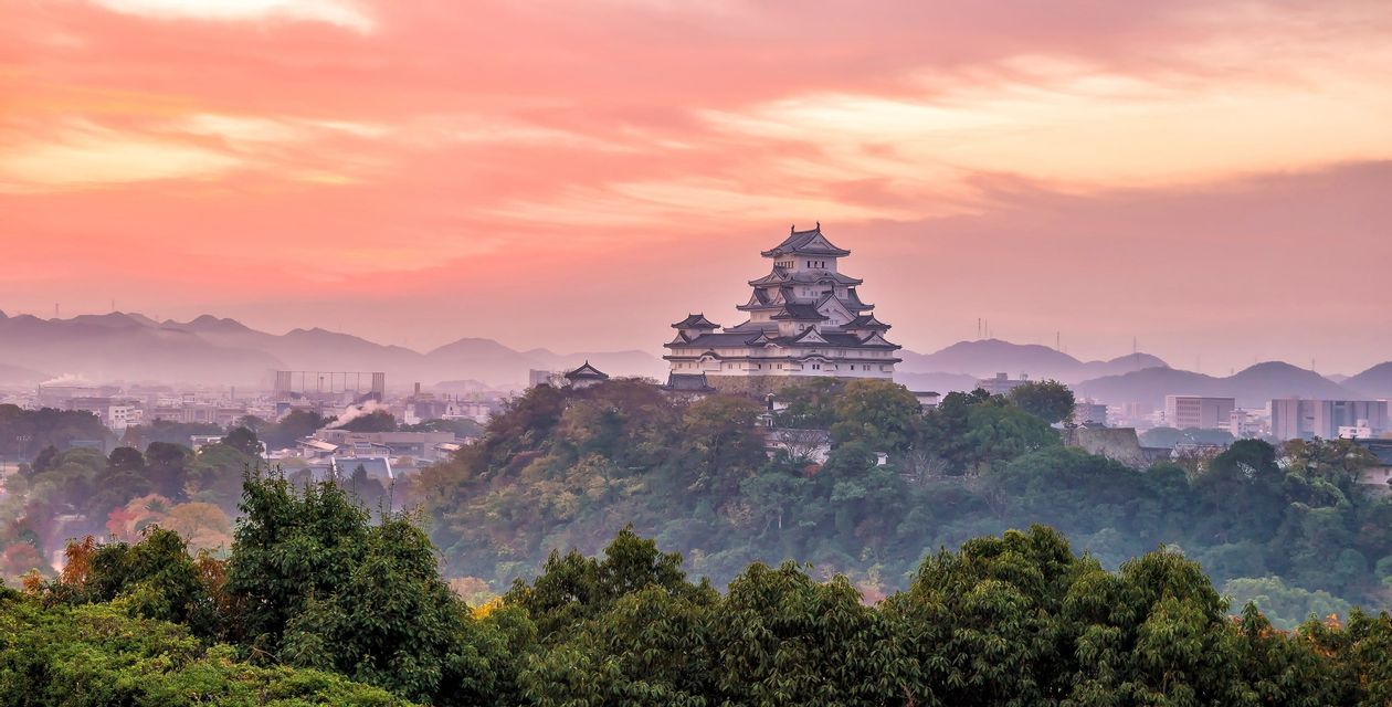 Un château japonais traditionnel sur une colline boisée surplombant une ville avec des montagnes brumeuses au coucher du soleil.