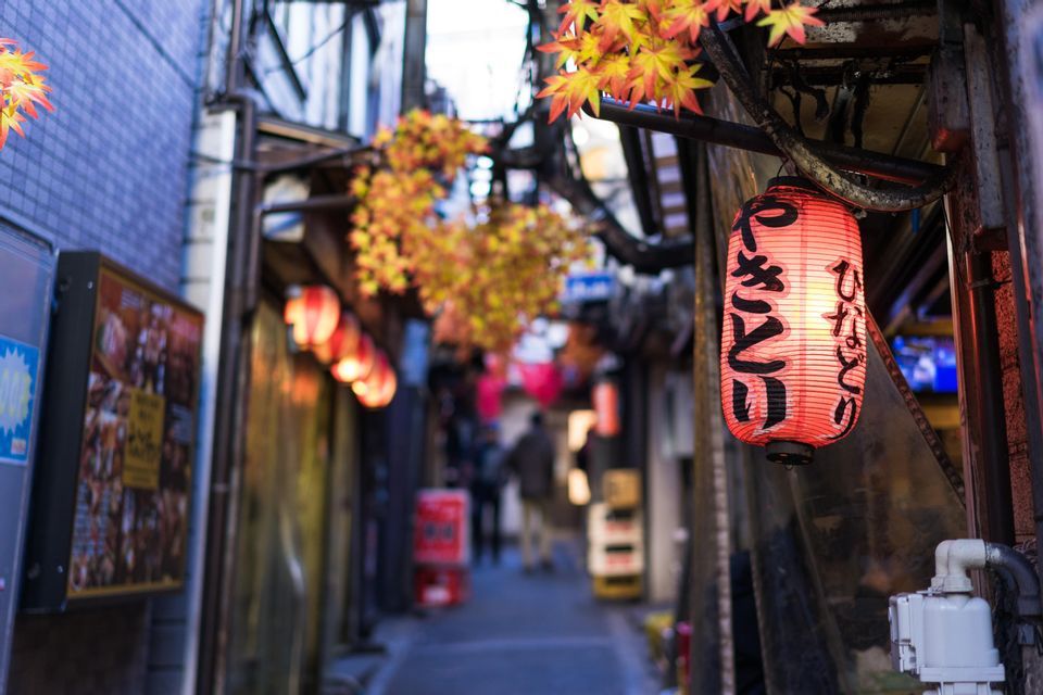 A close-up of an illuminated red Japanese paper lantern with calligraphy, hanging in a narrow city alley decorated with autumn leaves.