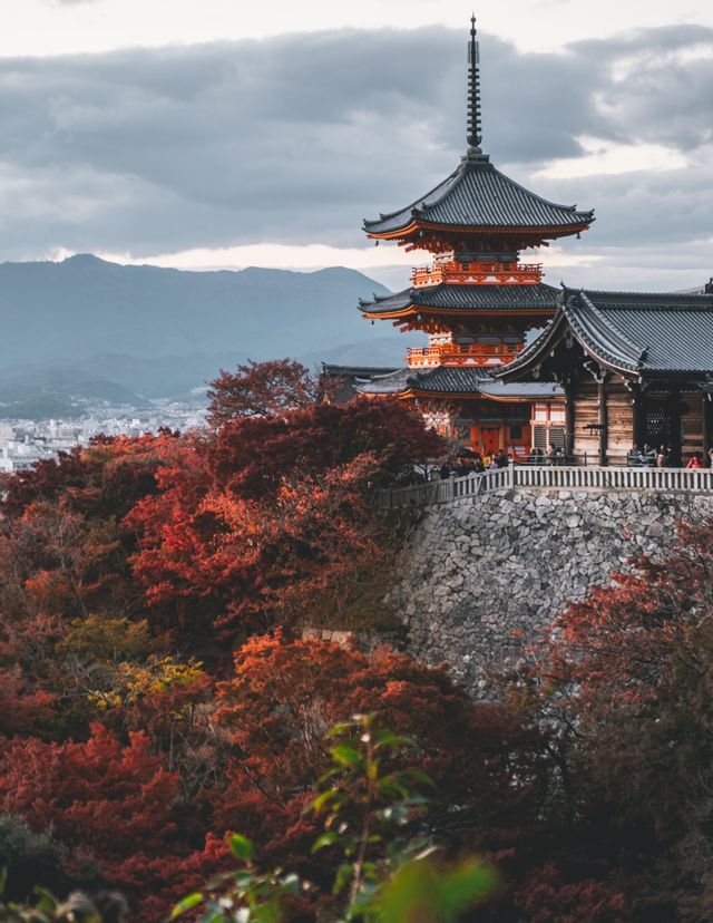 A red and black Japanese pagoda overlooks a dense forest with red autumn foliage, with mountains in the background under a cloudy sky.