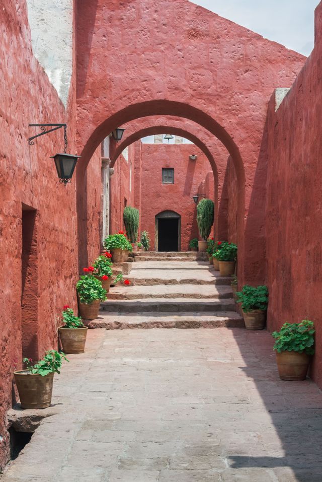 Un chemin de pierre avec des escaliers monte à travers un étroit couloir aux murs rouges et aux arches, bordé de géraniums en pot.