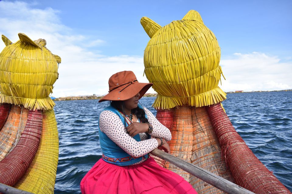 Una mujer con ropa tradicional colorida se sienta en una embarcación decorativa de totora en un lago grande bajo un cielo parcialmente nublado.