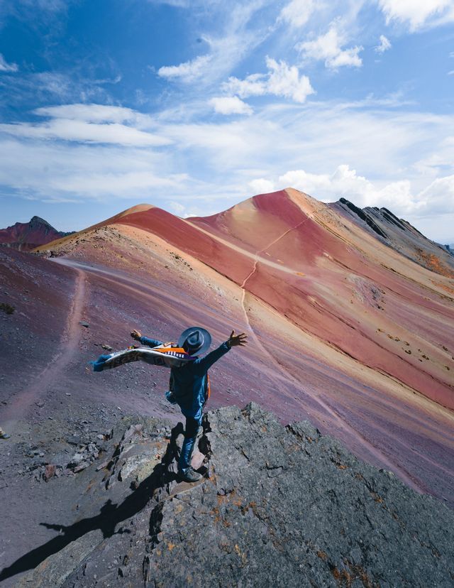 Une personne coiffée d'un chapeau se tient sur un promontoire rocheux, les bras tendus, dominant une chaîne de montagnes colorées et striées sous un ciel bleu.