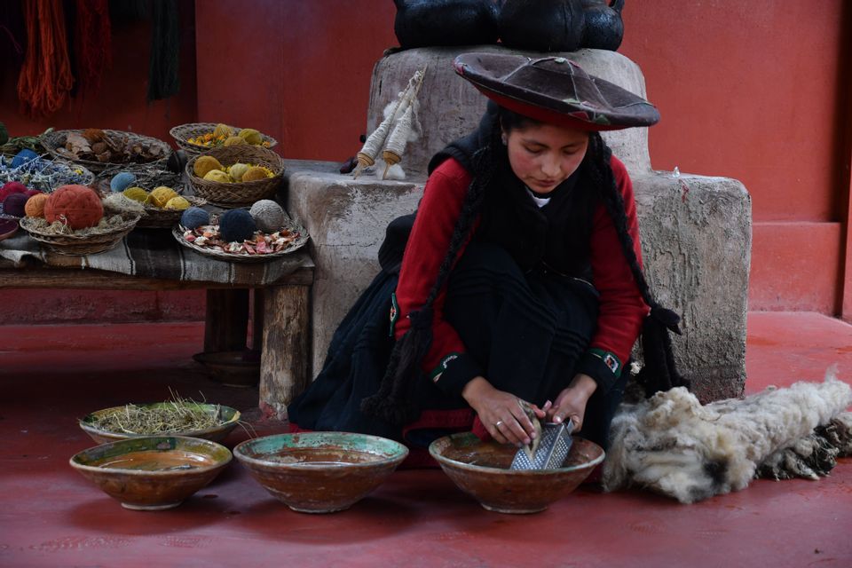 A woman in traditional clothing grates a root into a bowl to make natural dye, surrounded by raw wool and colorful yarn.
