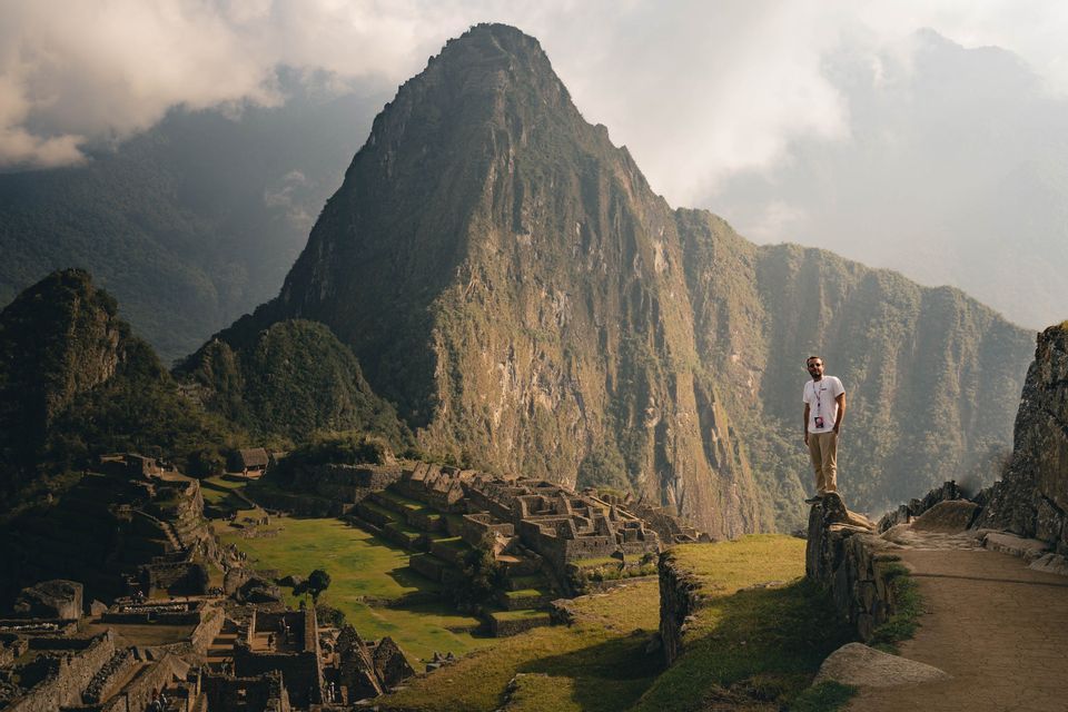 Un homme en t-shirt blanc se tient sur un mur de pierre dominant des ruines antiques et une grande montagne verte sous un ciel nuageux.