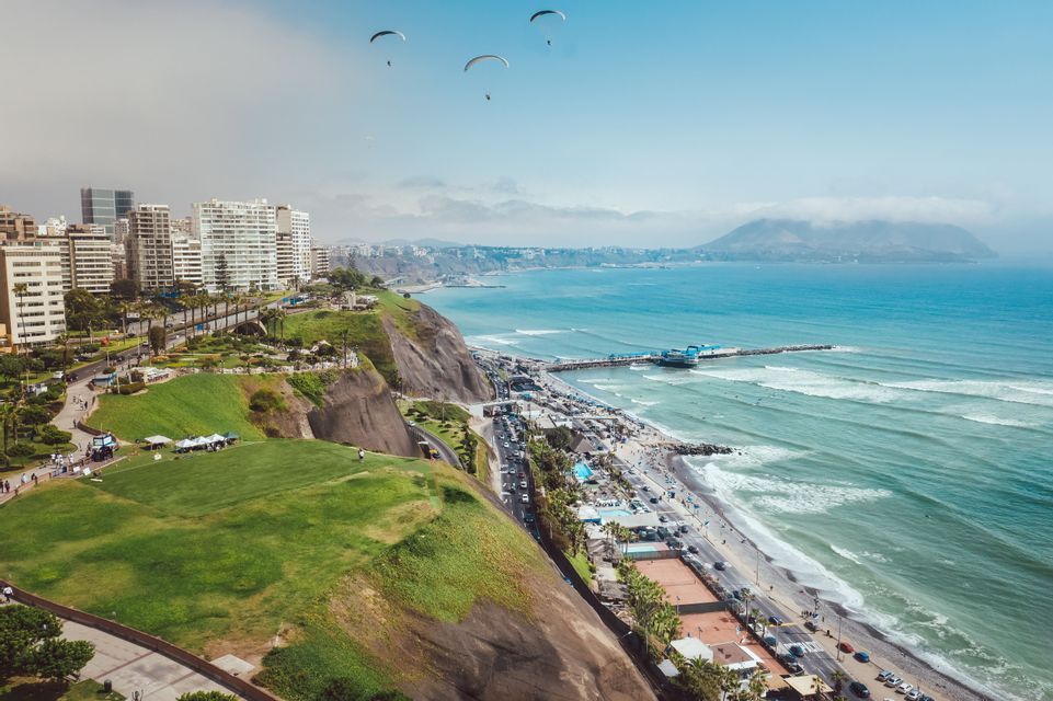 Una vista aérea de una ciudad costera en acantilados, con varios parapentistas volando sobre el océano y un muelle.