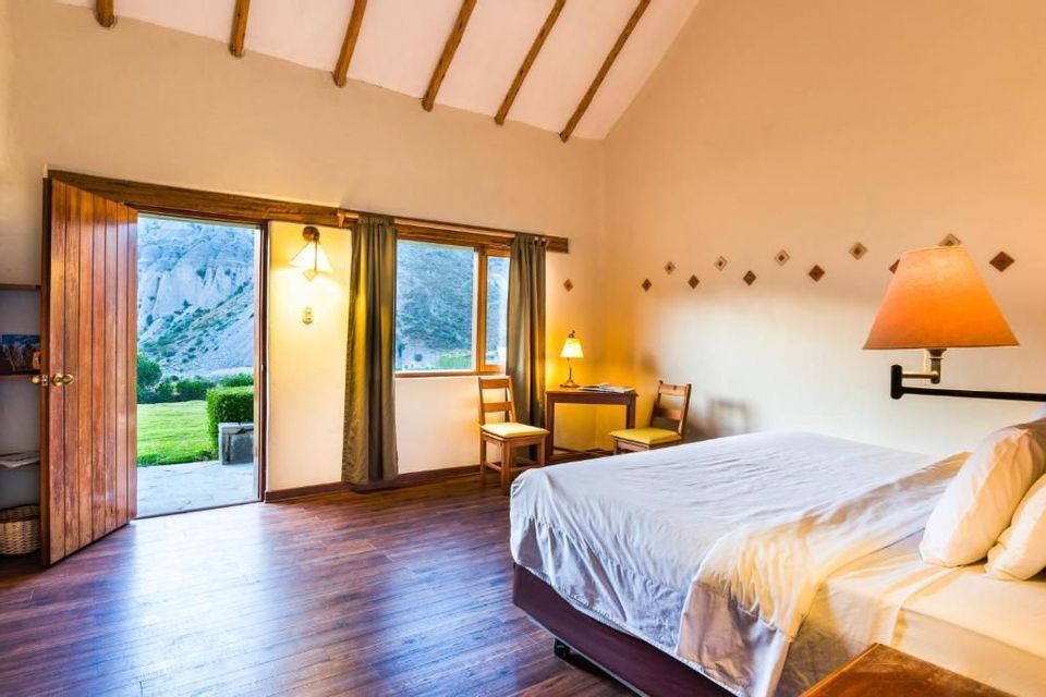 Una camera d'albergo con letto rifatto e pavimenti in legno ha porte aperte che rivelano una vista su un prato e montagne rocciose.