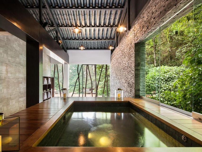 Una piscina interna in una stanza con un patio in legno, una parete in pietra a vista e grandi finestre che si affacciano su una foresta lussureggiante.