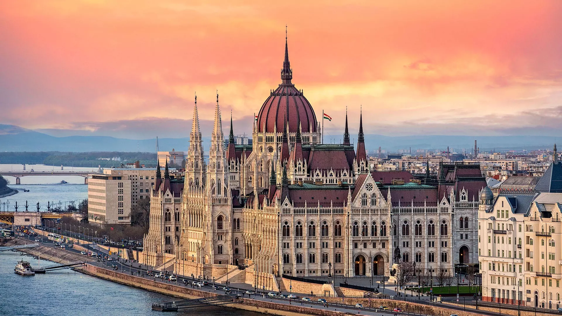 Un edificio del parlamento ornamentado con una gran cúpula se alza a la orilla de un río en el horizonte de una ciudad al atardecer.
