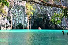 A small boat with people is rowing inside a large cave entrance, coming from a turquoise lagoon surrounded by lush green cliffs.