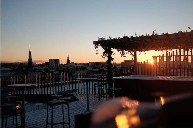 An empty panoramic terrace with tables and stools overlooks an urban landscape with a church bell tower during a fiery sunset.