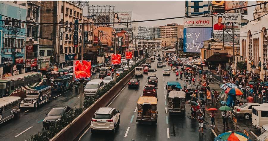 A bird's-eye view of a busy city street with cars, jeepneys, and buses on a wet road, and pedestrians on the sidewalks.