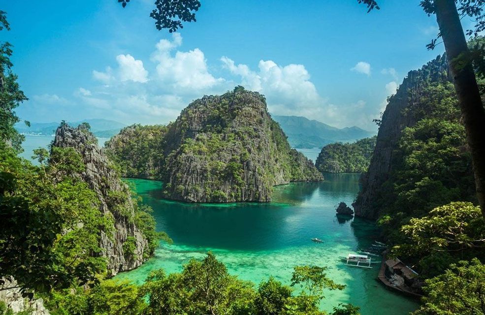 An aerial view shows a sunken wreck and a boat in a clear turquoise lagoon, surrounded by lush, rocky islands under a blue sky.