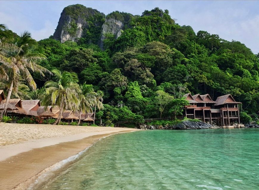 Thatched-roof bungalows and palm trees line a sandy beach next to clear turquoise water, backed by a lush, jungle-covered mountain.