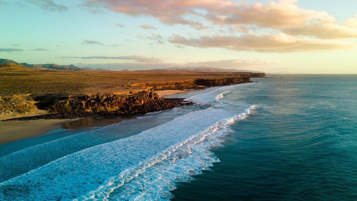 Vue aérienne de vagues turquoise se brisant le long d'un littoral accidenté et aride avec des montagnes lointaines sous un ciel coloré.