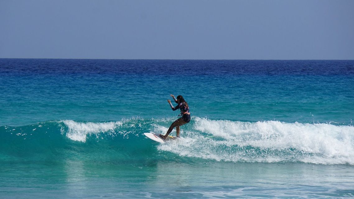 Una donna in muta fa surf su una tavola bianca, cavalcando un'onda turchese nell'oceano blu sotto un cielo sereno.