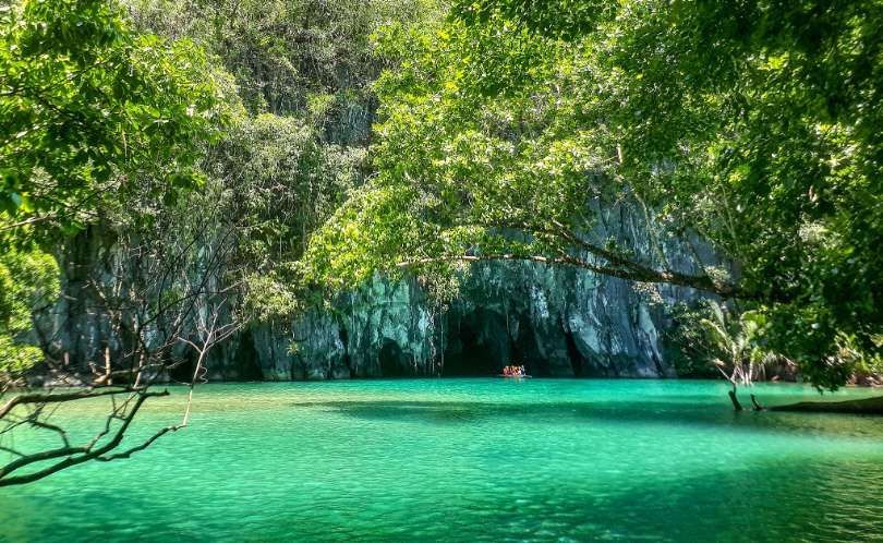 Un viaggio di gruppo WeRoad in barca naviga su acque turchesi verso l'apertura di una grotta in una grande scogliera rocciosa ricoperta di giungla.