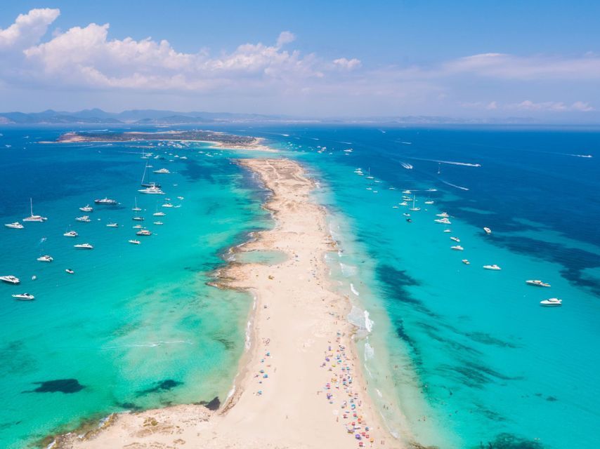 Una vista aerea di una lunga lingua di sabbia che si estende in un mare turchese, con molte barche ancorate e persone sulla spiaggia.