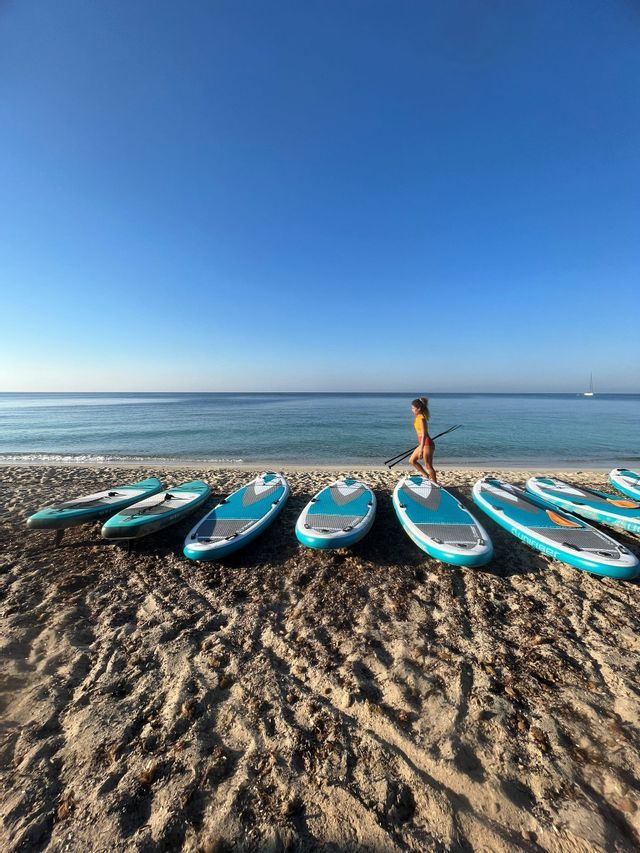 Una mujer con una pala pasa junto a varias tablas de paddle surf alineadas en una playa de arena junto al mar.
