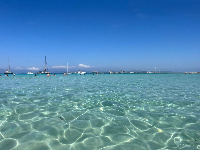Persone che nuotano in acque limpide e turchesi, con numerose barche a vela e yacht ancorati in lontananza sotto un cielo azzurro brillante.