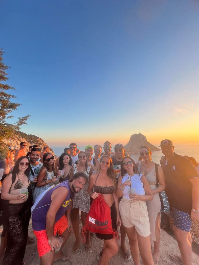 Un viaje en grupo de WeRoad posa para una foto en un acantilado con vistas al mar y a una isla rocosa al atardecer.