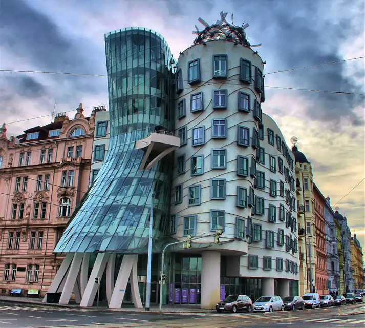 Un edificio moderno con una torre di vetro attorcigliata e una torre di cemento con finestre ondulate, su un angolo di strada cittadina.