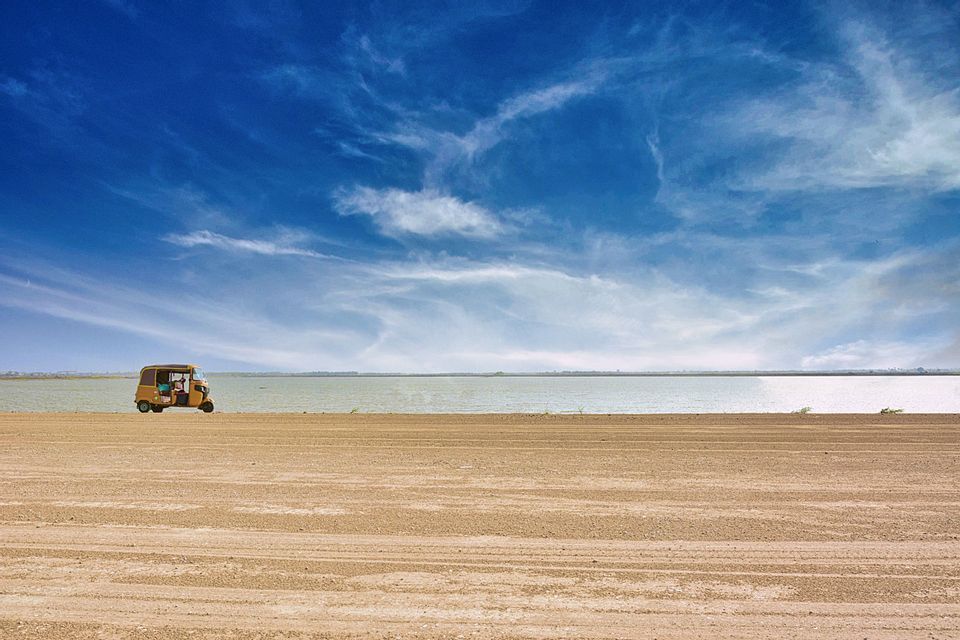Un tuk-tuk giallo parcheggiato su una vasta spiaggia sabbiosa accanto a uno specchio d'acqua sotto un ampio cielo blu con nuvole striate.