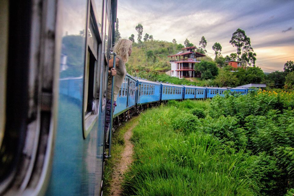 Una donna bionda si sporge da un treno blu che si snoda attraverso un paesaggio lussureggiante e verde, tra le colline.