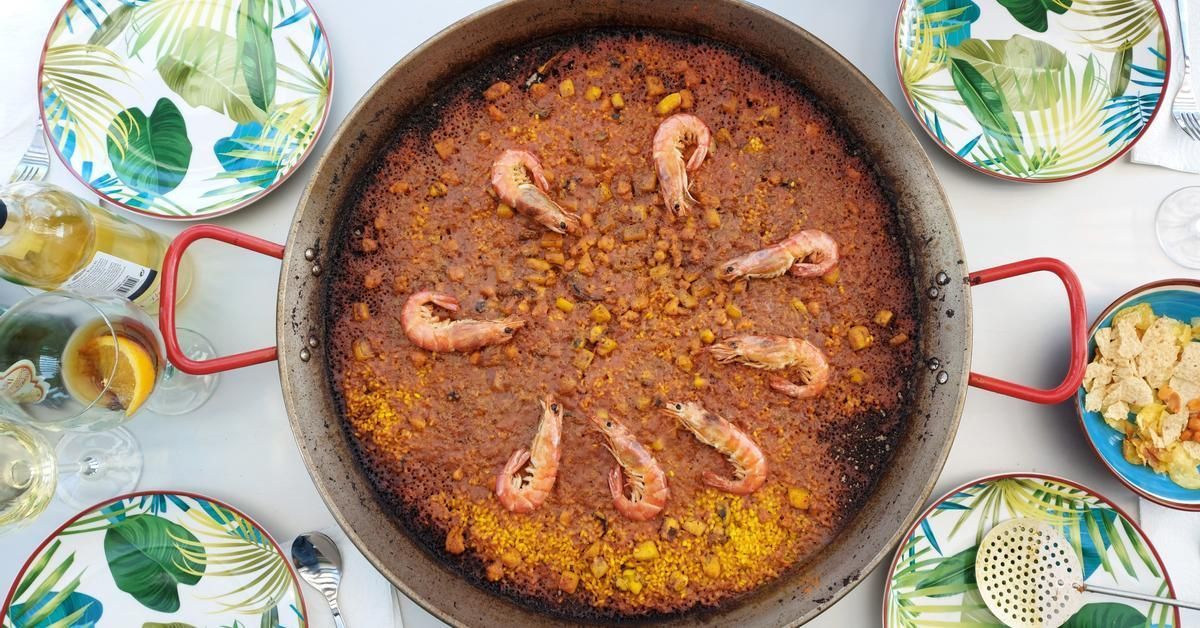 A top-down view of a large paella pan filled with rice and prawns, surrounded by leaf-patterned plates and glasses.