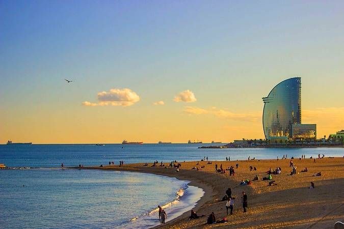 People relaxing on a sandy beach during sunset, with a large, sail-shaped glass building next to the sea in the background.