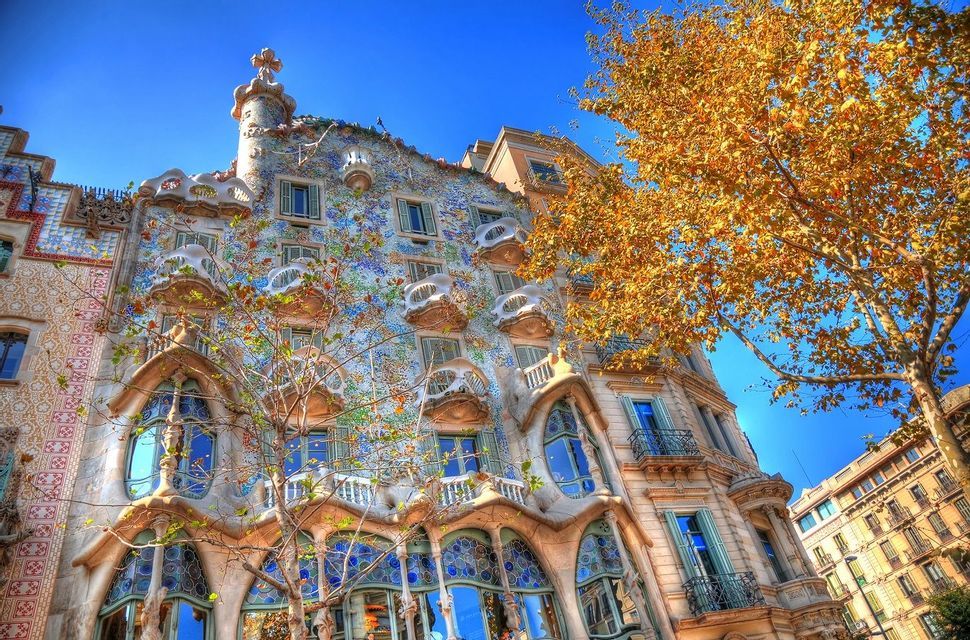 Una ripresa dal basso di un edificio con una facciata a mosaico colorata e balconi scultorei, accanto a un albero con foglie gialle contro un cielo blu.
