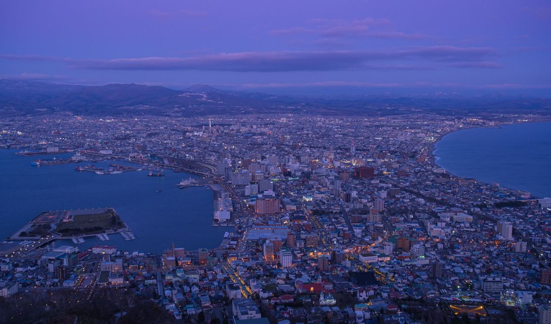 Una vista aerea di una vasta città costiera con strade ed edifici illuminati al crepuscolo, con montagne sullo sfondo.