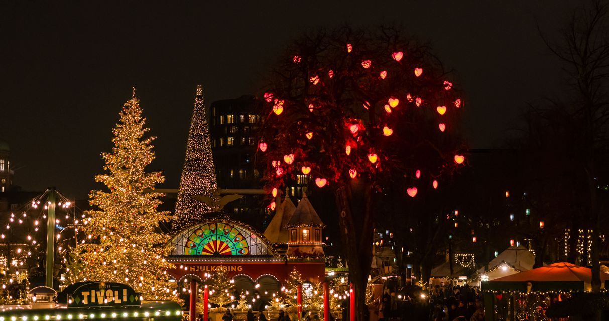 Ein festlicher Freizeitpark bei Nacht, beleuchtet von großen Weihnachtsbäumen und einem weiteren Baum mit leuchtend roten, herzförmigen Lichtern.