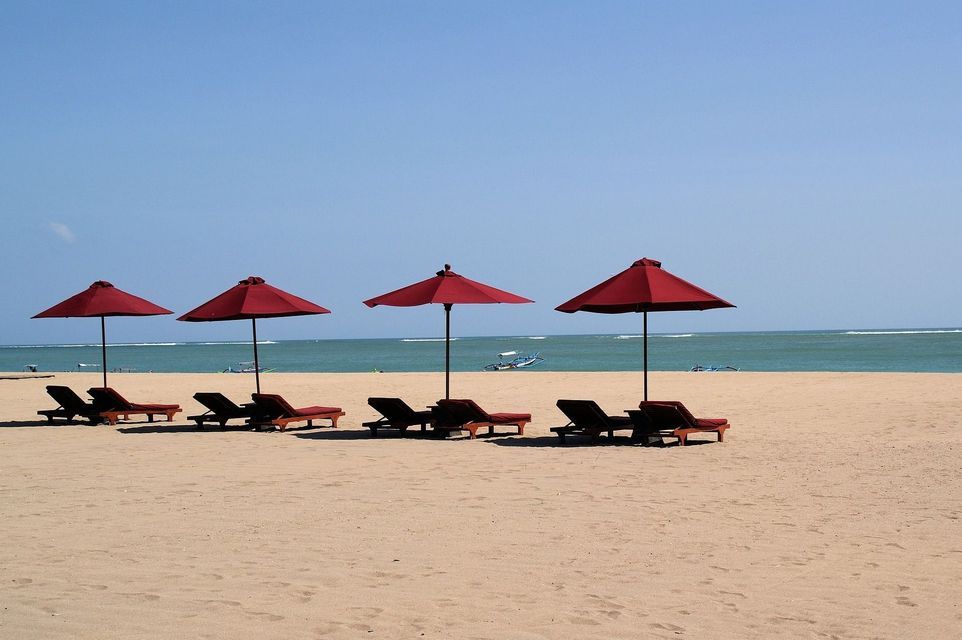 Une rangée de parasols rouges et de transats vides sur une plage de sable fin face à l'océan, sous un ciel dégagé.