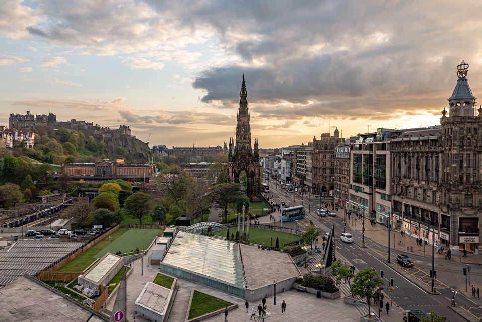 An elevated view of a historic cityscape at sunset, with a castle on a hill and an ornate monument.