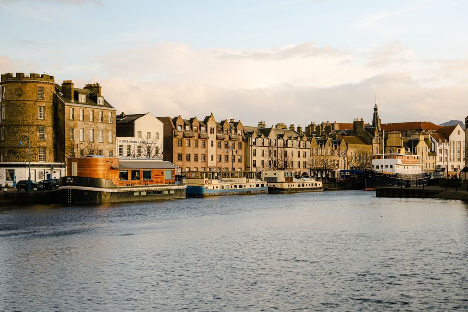 Historic city buildings and moored houseboats line a calm river under a cloudy sky at sunset.