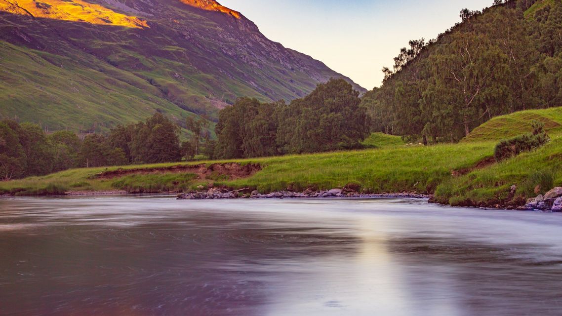 A river with smoothed water flows past a green hillside, with a mountain peak lit by golden light.