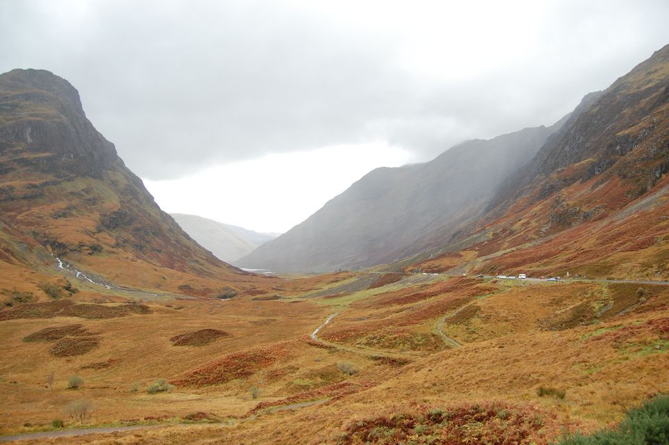 Une large vallée montagneuse avec des pentes couvertes de feuillage d'automne marron et orange sous un ciel couvert.