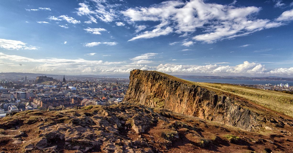 A view from a rocky cliff overlooking a vast city skyline and a body of water under a partly cloudy blue sky.