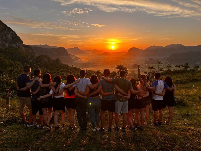 A WeRoad group trip stands with arms around each other, watching the sun set over a misty mountain valley from a grassy viewpoint.