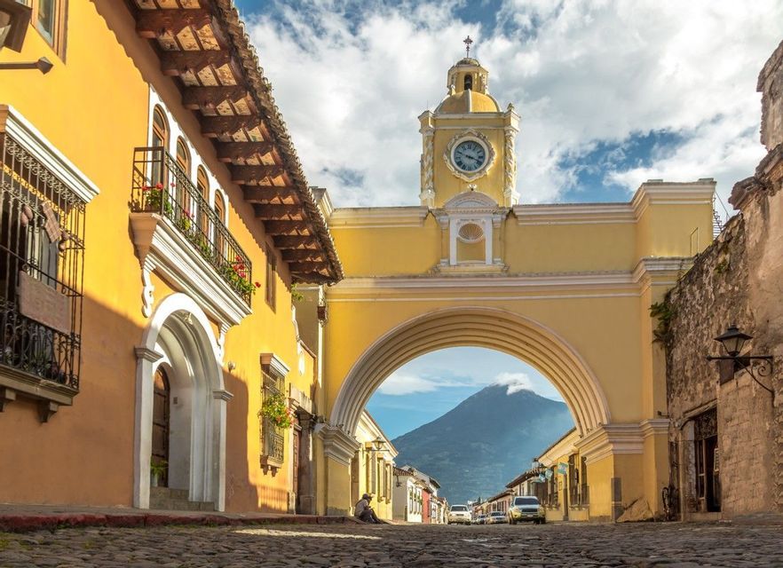 Un arco giallo della torre dell'orologio si estende su una strada di ciottoli, incorniciando un vulcano lontano sotto un cielo parzialmente nuvoloso.