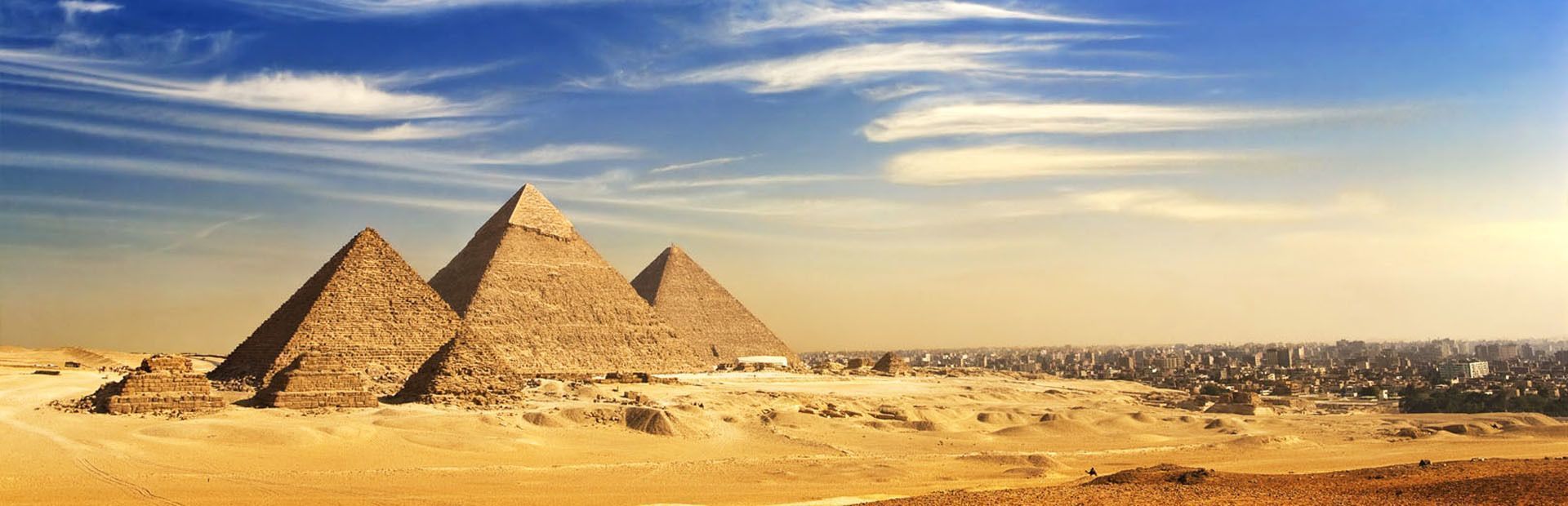 Una vista panorámica de tres grandes pirámides en un desierto arenoso con una ciudad en expansión en la distancia bajo un cielo azul y nublado.