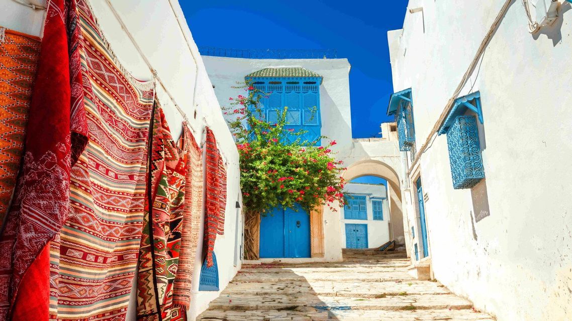 Uno stretto vicolo acciottolato costeggiato da edifici imbiancati a calce, porte blu vivaci e tappeti colorati appesi a una parete sotto un cielo soleggiato.