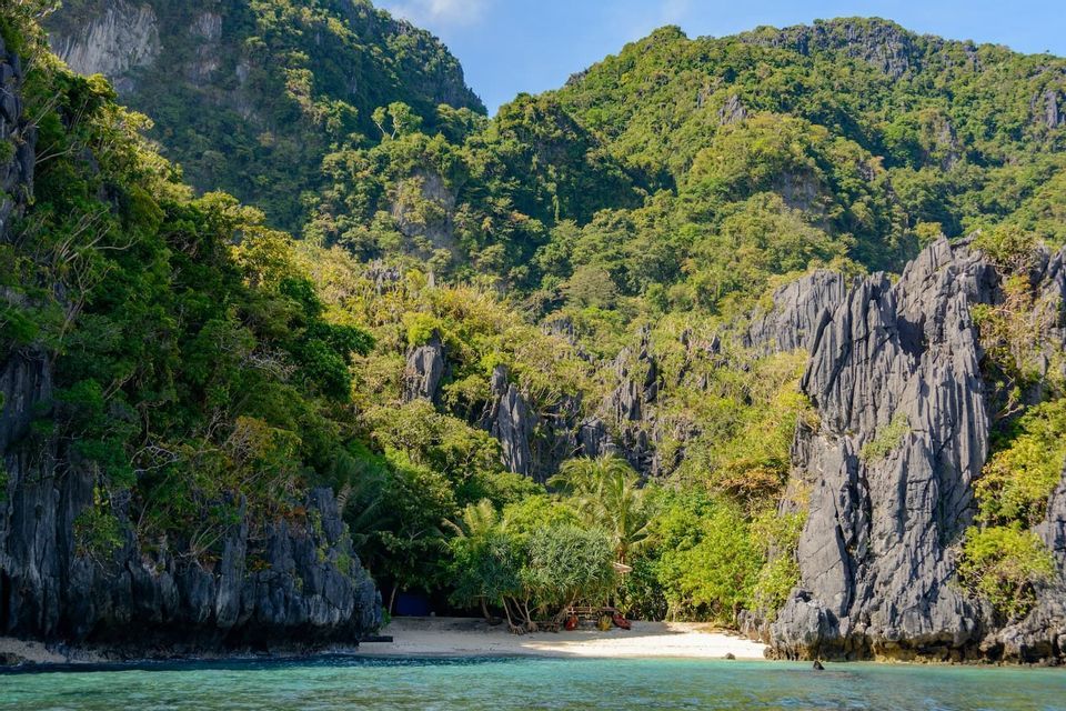 A hidden cove with a white sand beach, surrounded by lush green mountains and dark, jagged cliffs, seen from the turquoise water.