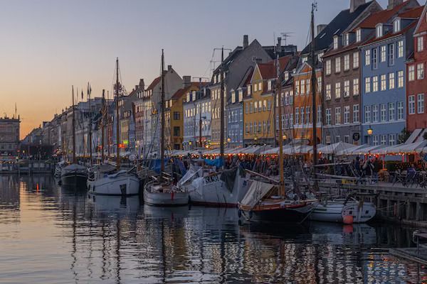 Des voiliers amarrés le long d'un canal, devant des bâtiments historiques colorés et des cafés en bord de quai, au crépuscule.
