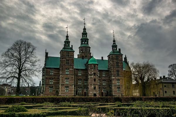 Un grand château de briques aux toits verts et aux flèches se dresse derrière un jardin soigné sous un ciel couvert.