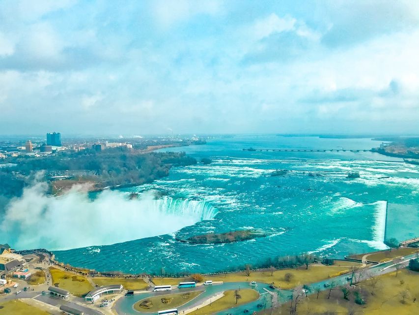 Eine Luftaufnahme eines großen, hufeisenförmigen Wasserfalls mit leuchtend türkisfarbenem Wasser, einem Aussichtsbereich und einer entfernten Stadtlandschaft.