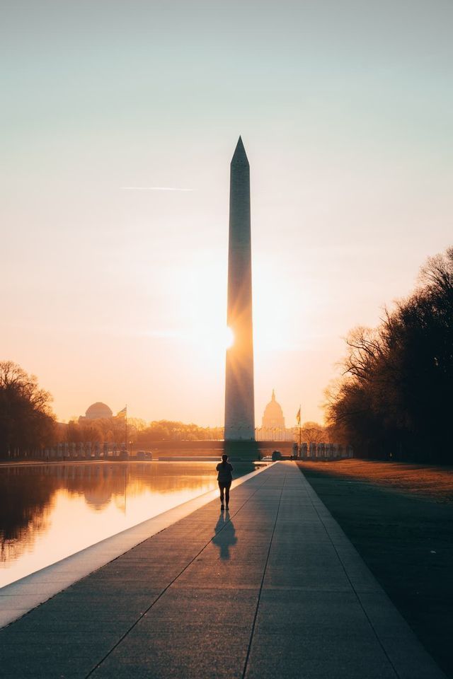Eine Person geht auf einem Pfad neben einem reflektierenden Becken auf einen hohen Obelisken zu, während die Sonne bei Sonnenaufgang dahinter aufleuchtet.