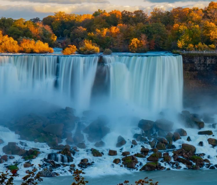 Ein breiter Wasserfall stürzt über einen felsigen Vorsprung in ein nebliges Becken, umgeben von einem Wald mit goldenen Herbstblättern.